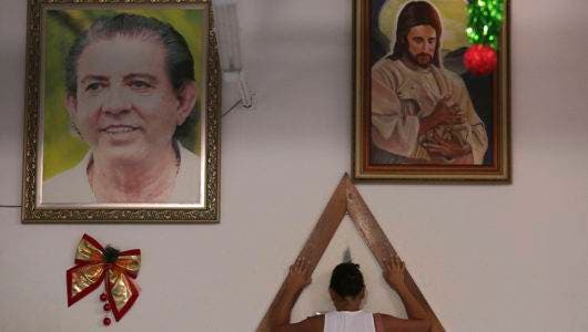 En esta fotografía del 4 de enero de 2019, una mujer coloca las manos sobre un triángulo de madera para rezar, ubicado entre cuadros del líder espiritual Joao Teixeira de Faria y de Jesucristo, en Abadiania, Brasil. (AP Foto/Eraldo Peres)
